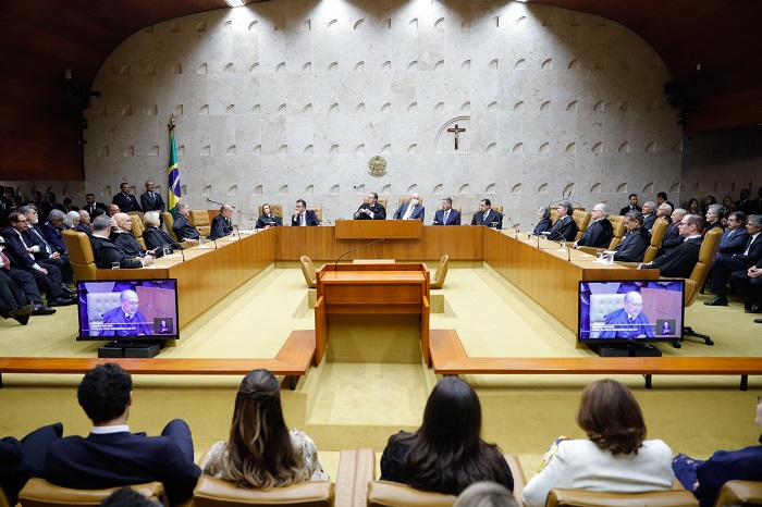 Foto de um plenário, composto por várias pessoas sentadas em poltronas de cor marrom claro. Ao centro, duas televisões. 