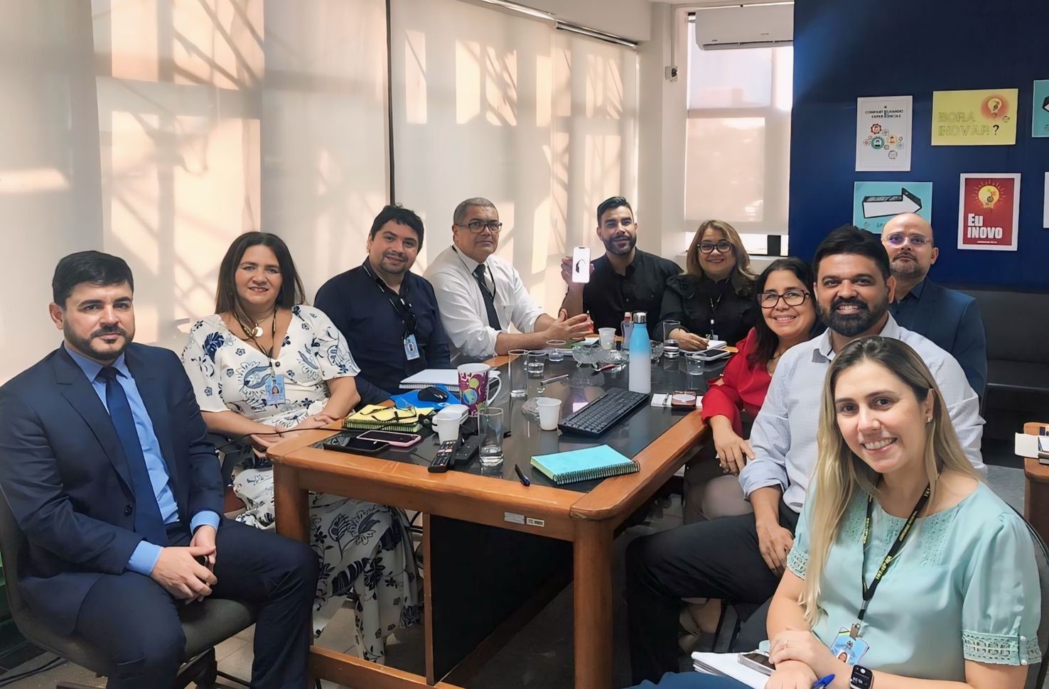 Várias pessoas, homes e mulheres, sentados em torno de uma mesa, posando para a foto. Eles estão dentro de uma sala.