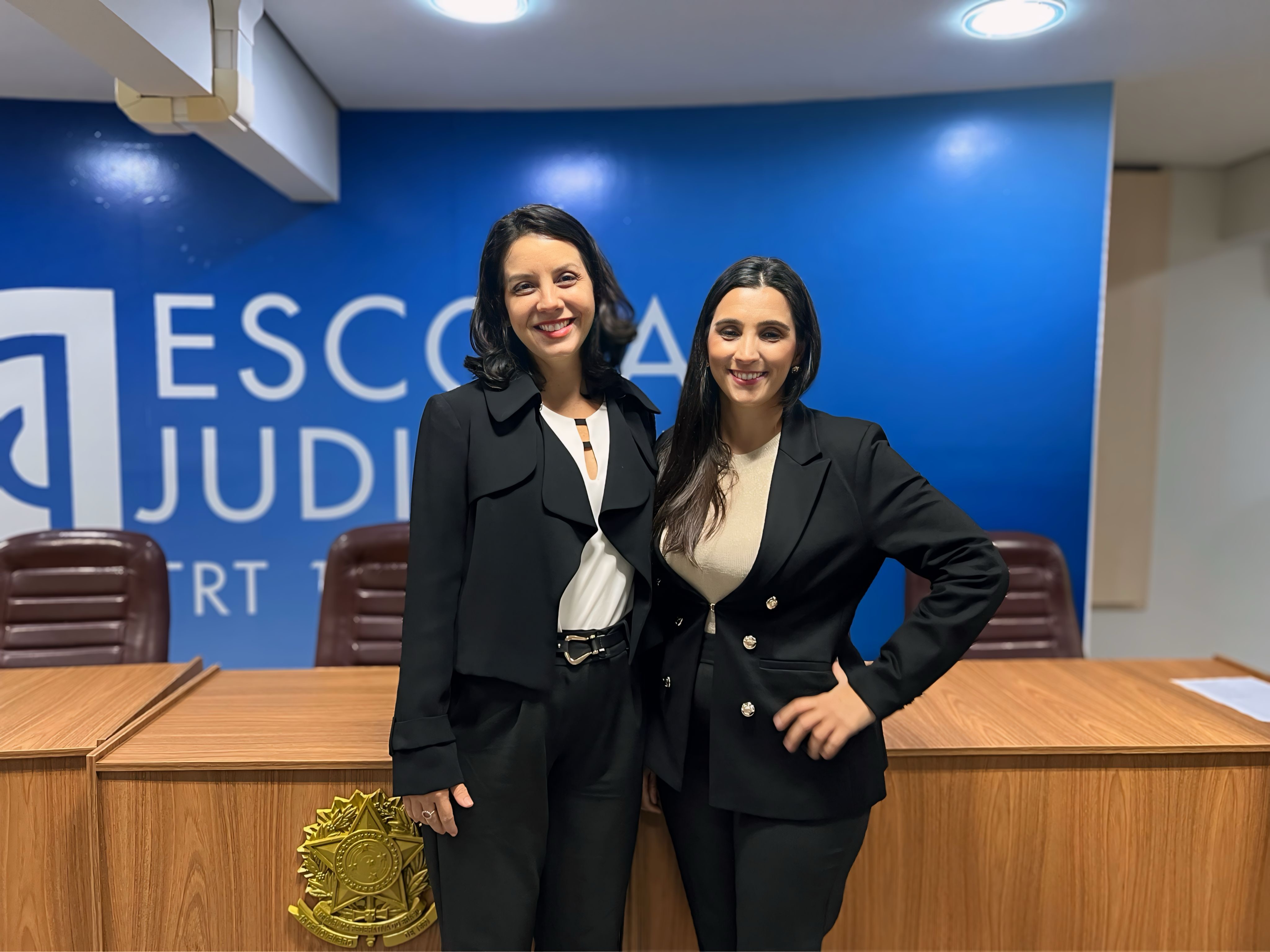 Duas pessoas posando para foto, dentro de uma sala. Ao fundo, parede azul escrita com letras brancas ESCOLA JUDICIAL.