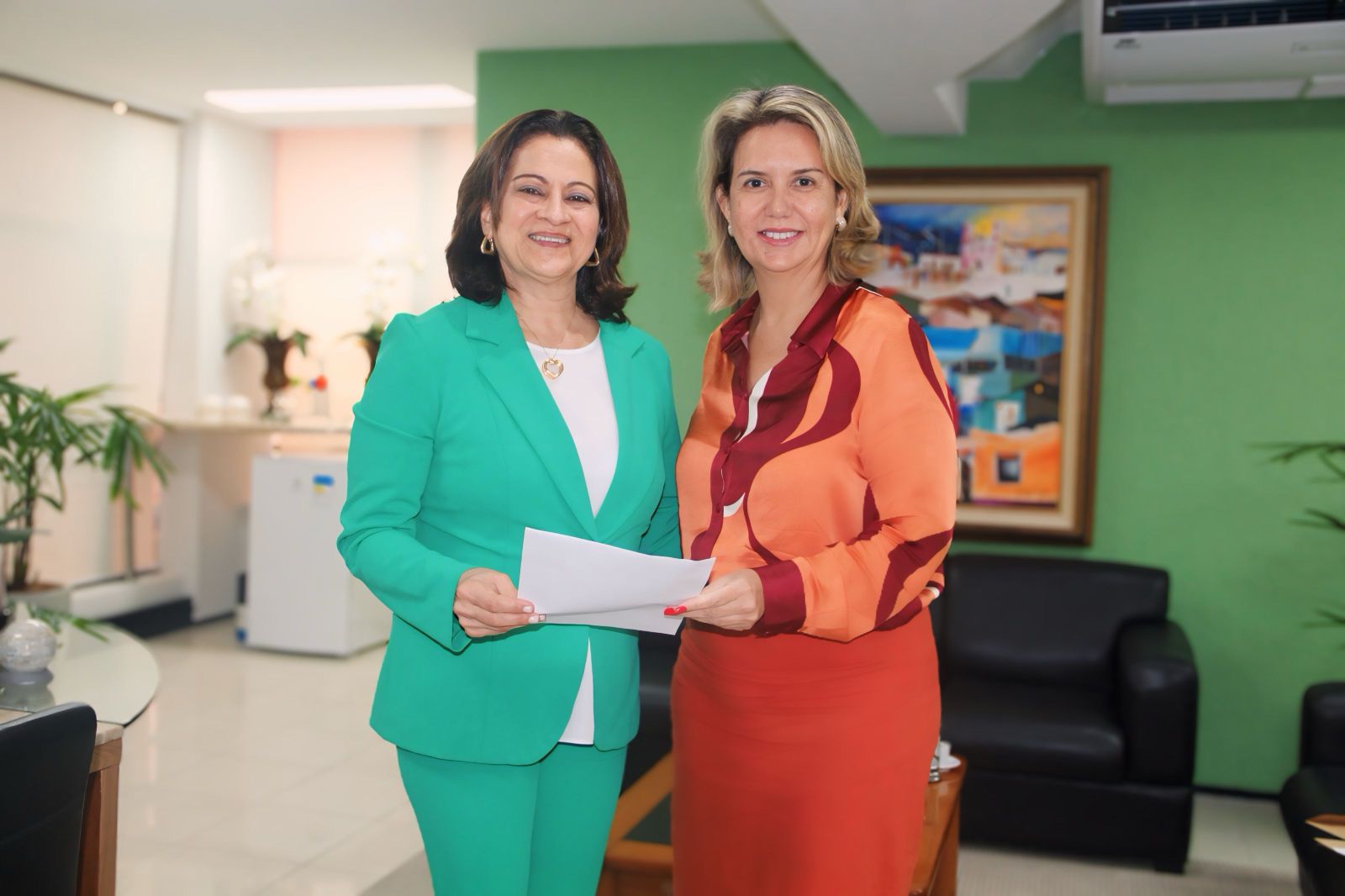 Duas mulheres posando para foto, dentro de uma sala com paredes no fundo esverdeadas. Elas seguram juntas uma folha de papel.