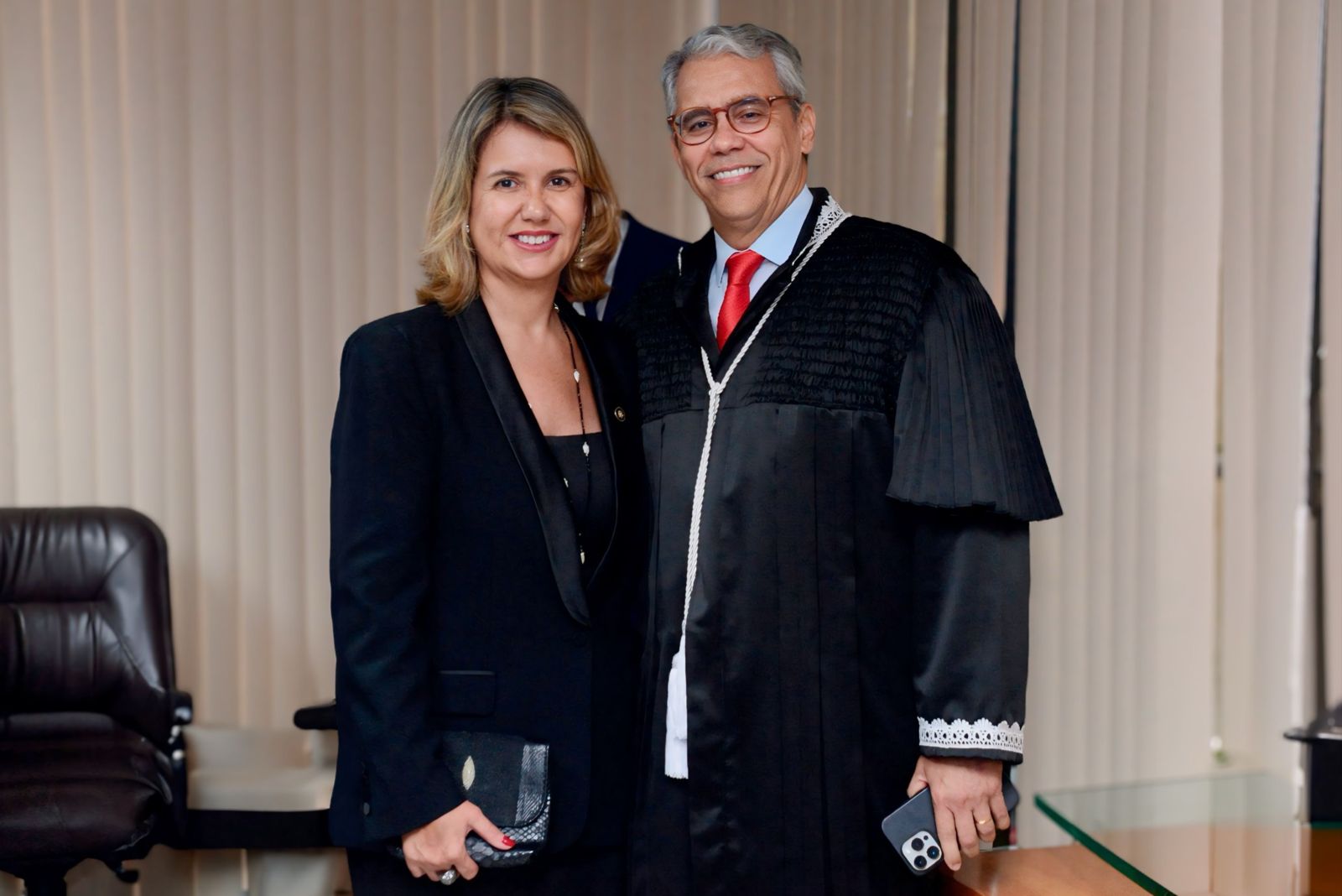 Duas pessoas posando para foto dentro de uma sala. Uma mulher, com vestido e blazer preto, segurando na mão uma bolsa preta, e um homem vestido com toga preta.