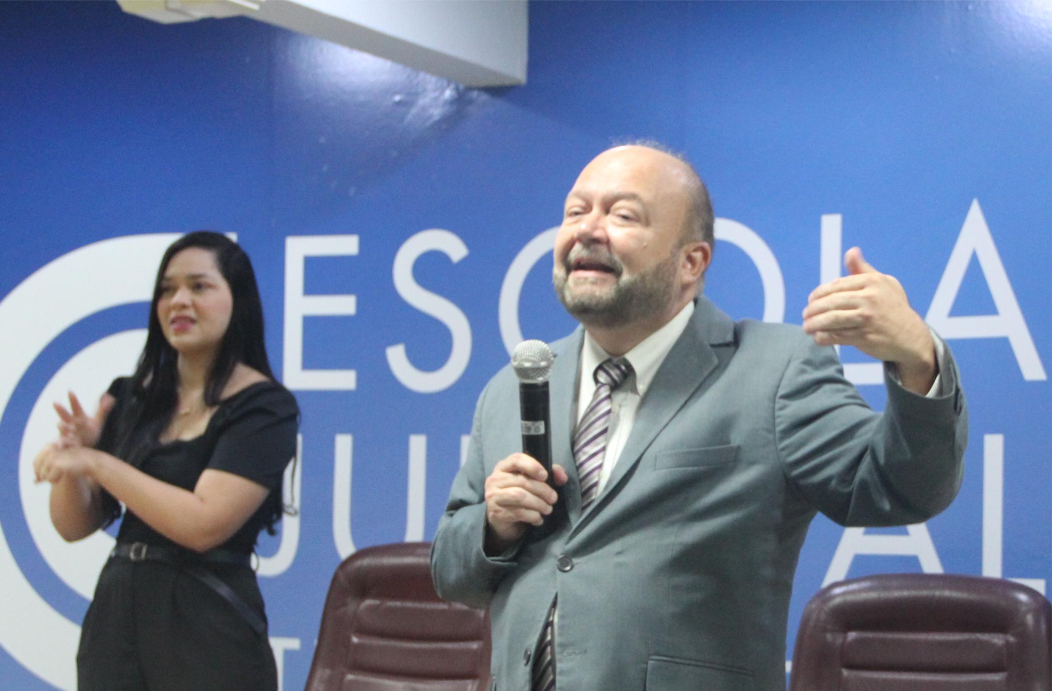 Foto de um homem branco e calvo, sorrindo e falando ao microfone, que segura com a mão direita. Ele usa terno cinza e gesticula com a mão esquerda. Em segundo plano, vê-se uma mulher branca de cabelos longos e pretos, usando roupa preta, gesticulando ao interpretar em LIBRAS a fala do homem. Ao fundo, vê-se parede azul com o texto ESCOLA JUDICIAL escrito em letras brancas, e os espaldares de duas cadeiras marrons.