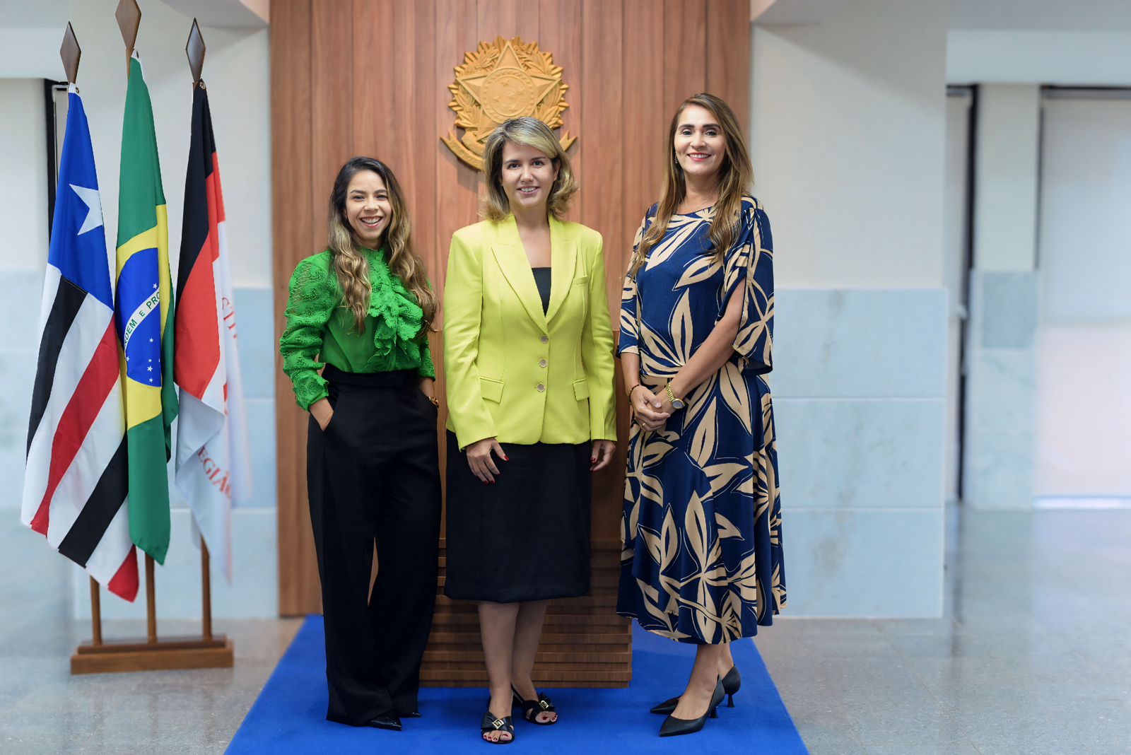 Três mulheres posando para foto. Ao fundo, banderias do Maranhão, Brasil e do TRT-16 hasteadas. 