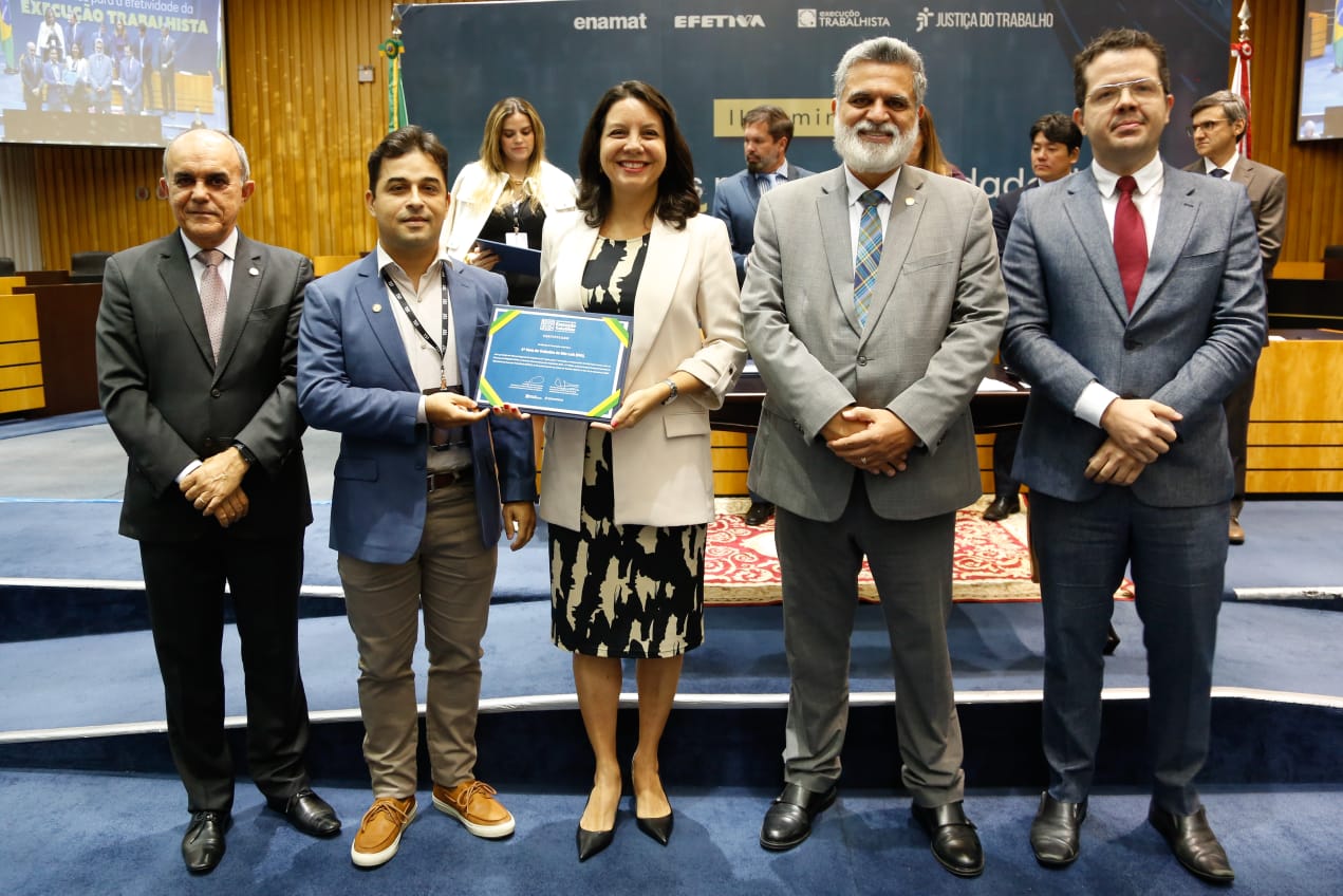 Cinco pessoas posam para uma foto dentro de um auditório. Quatro homens e uma mulher estão presentes, com a mulher localizada ao centro. A mulher e o homem à sua esquerda seguram uma placa comemorativa.