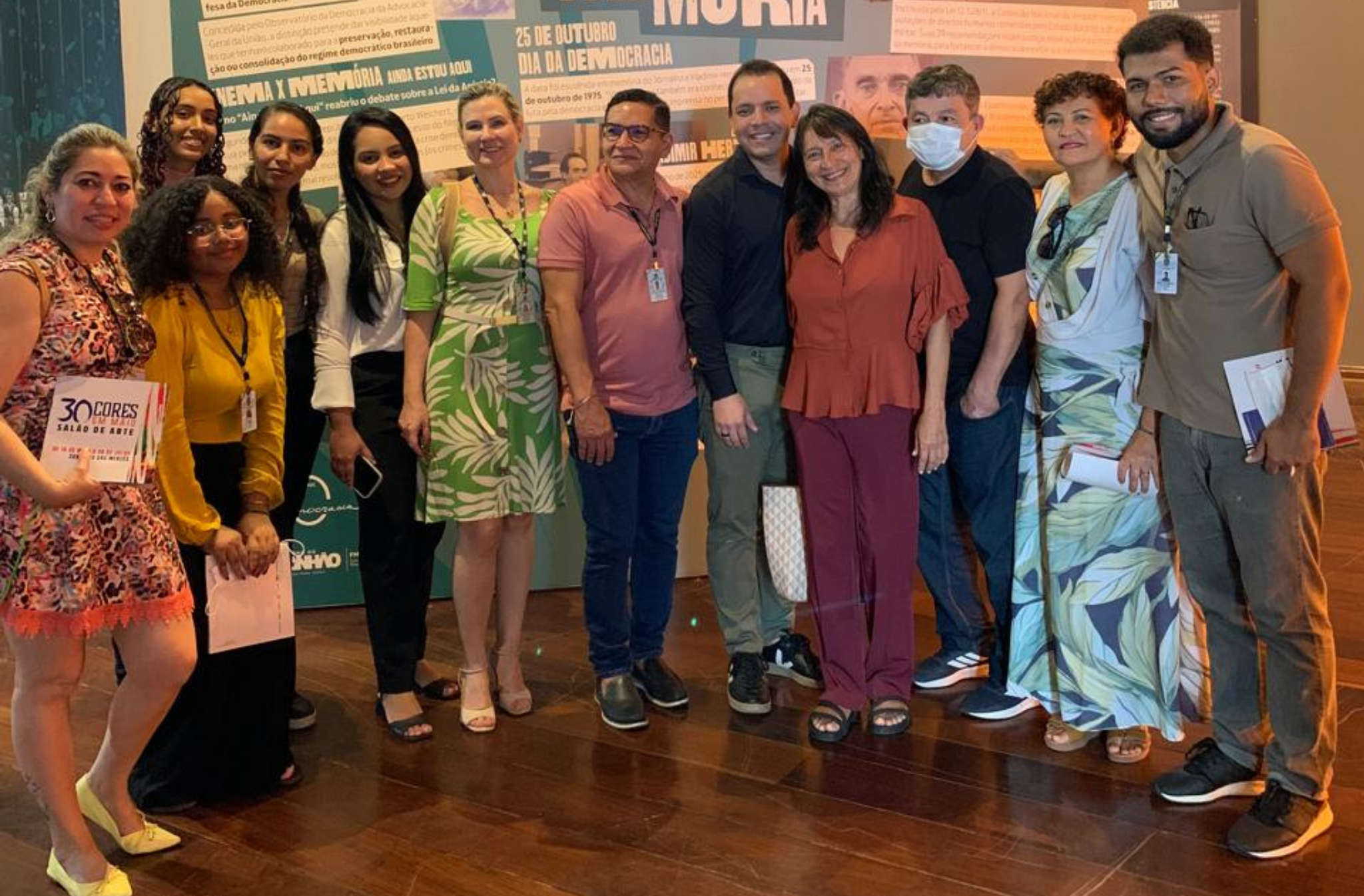 A imagem mostra uma foto de grupo em frente a um painel informativo sobre a "Preservação da Memória". As pessoas estão vestidAs com roupas casuais e alguns estão sorrindo para a câmera. O fundo é um espaço interno com piso de madeira.