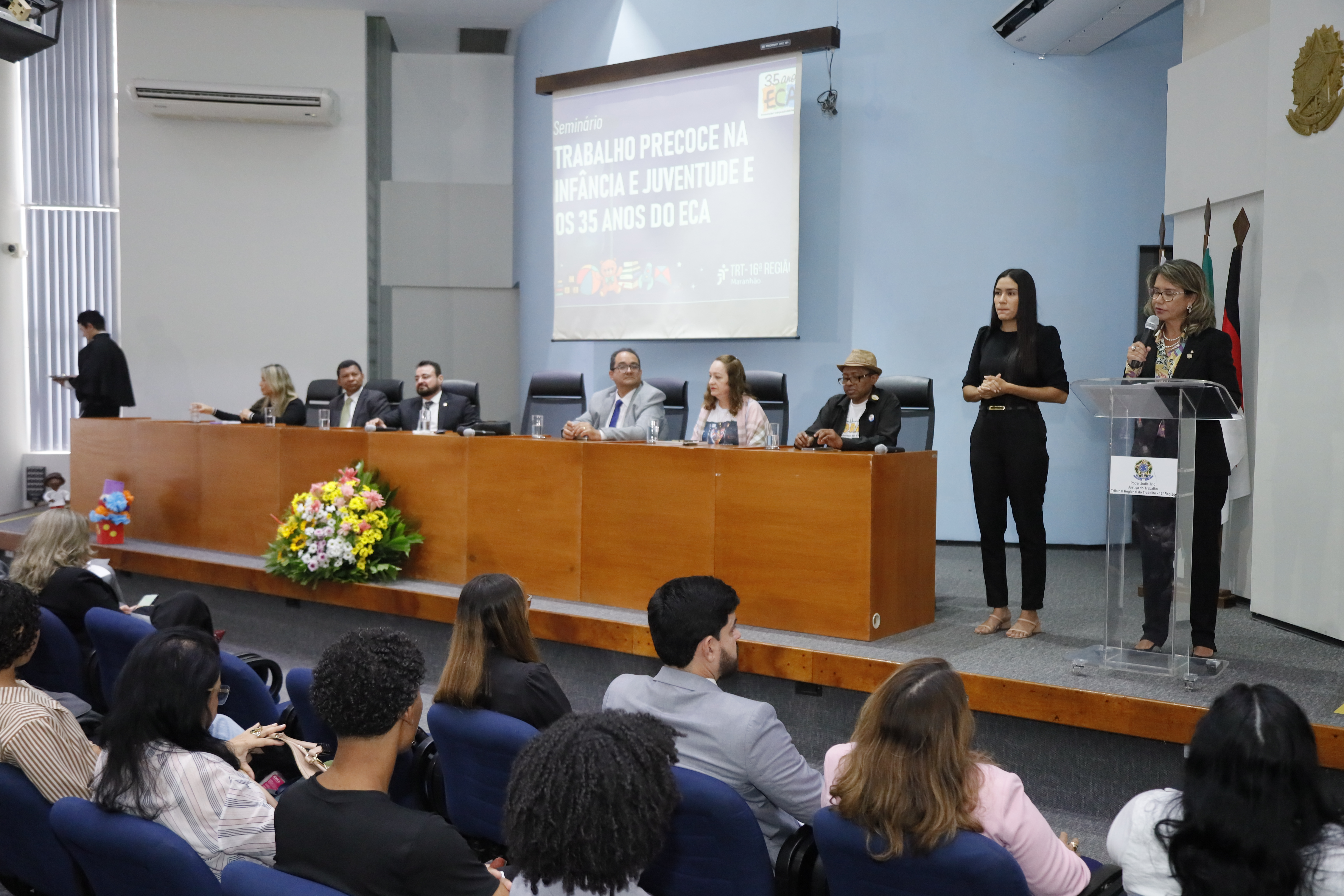 Foto mostra à direita mulher clara com roupa preta, atrás de um pulpito falando ao microfone. A sua direita, posiciona-se mulher com roupa preta, interprete de libras. A direita desta, mesa de honra com a presença de seis pessoas sentadas.