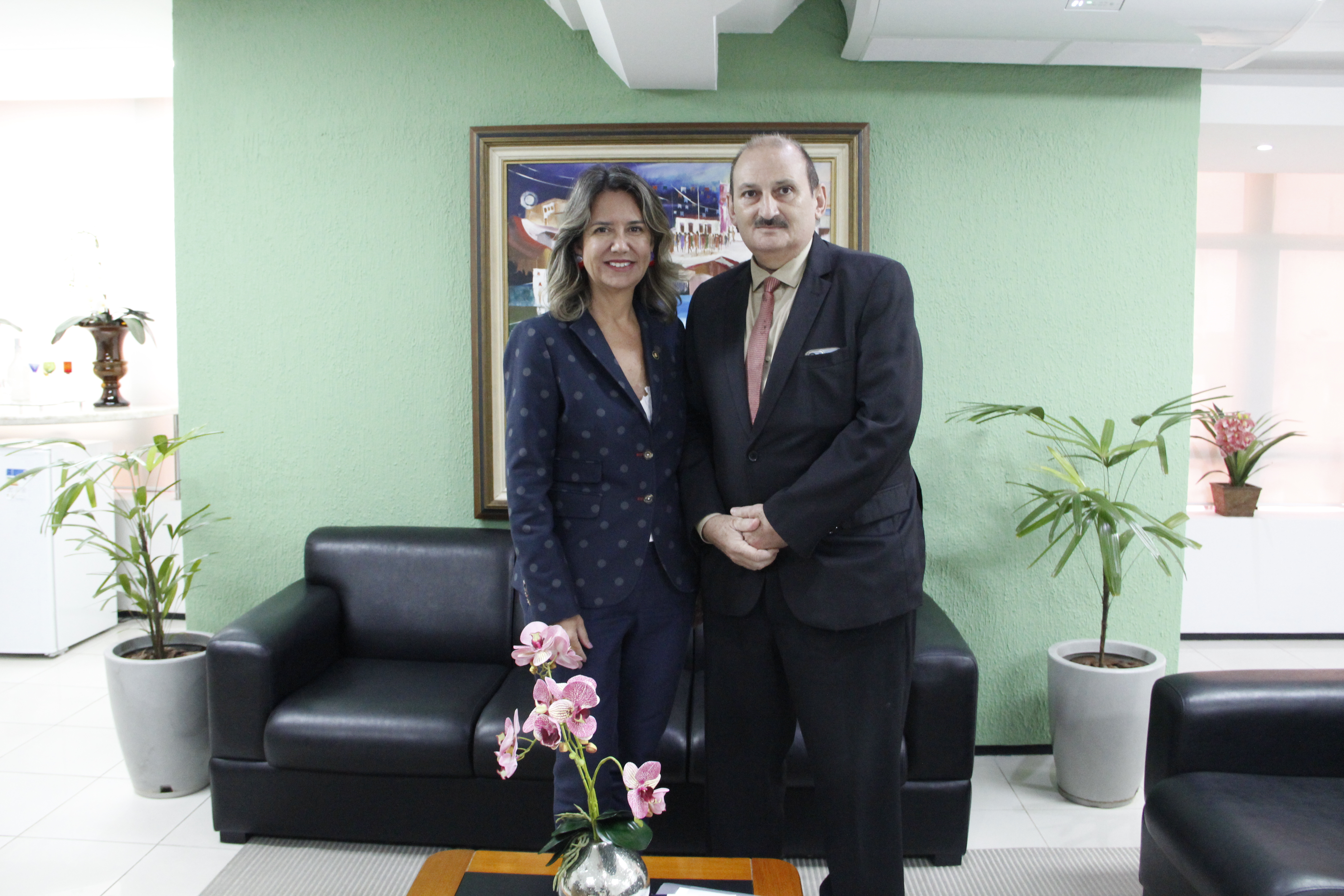 Foto mostra uma mulher e um homem. Ambos estão de pé. Ao fundo um ambiente de gabinete com sofá, quadros na paredes.