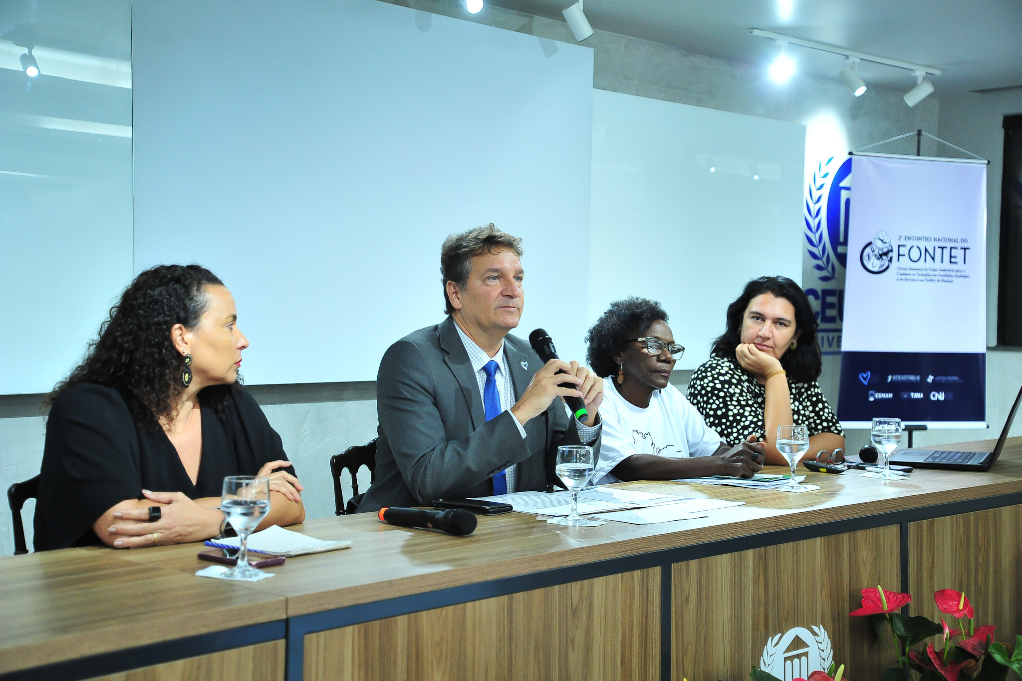 A imagem mostra quatro pessoas sentadas a uma mesa. Da esquerda para a direita, estão: uma mulher de cabelo preto; um homem de terno, usando um microfone; uma mulher negra, com cabelo curto e blusa clara; e uma mulher com cabelo preto. A mesa tem copos de água, microfones e papéis