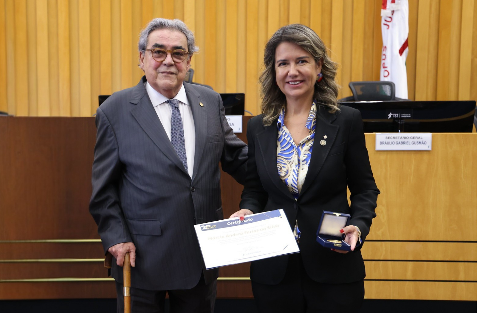 Foto mostra um homem e uma mulher segurando um certificado e uma medalha. Na parte do fundo, imagem de uma parede amaderada.