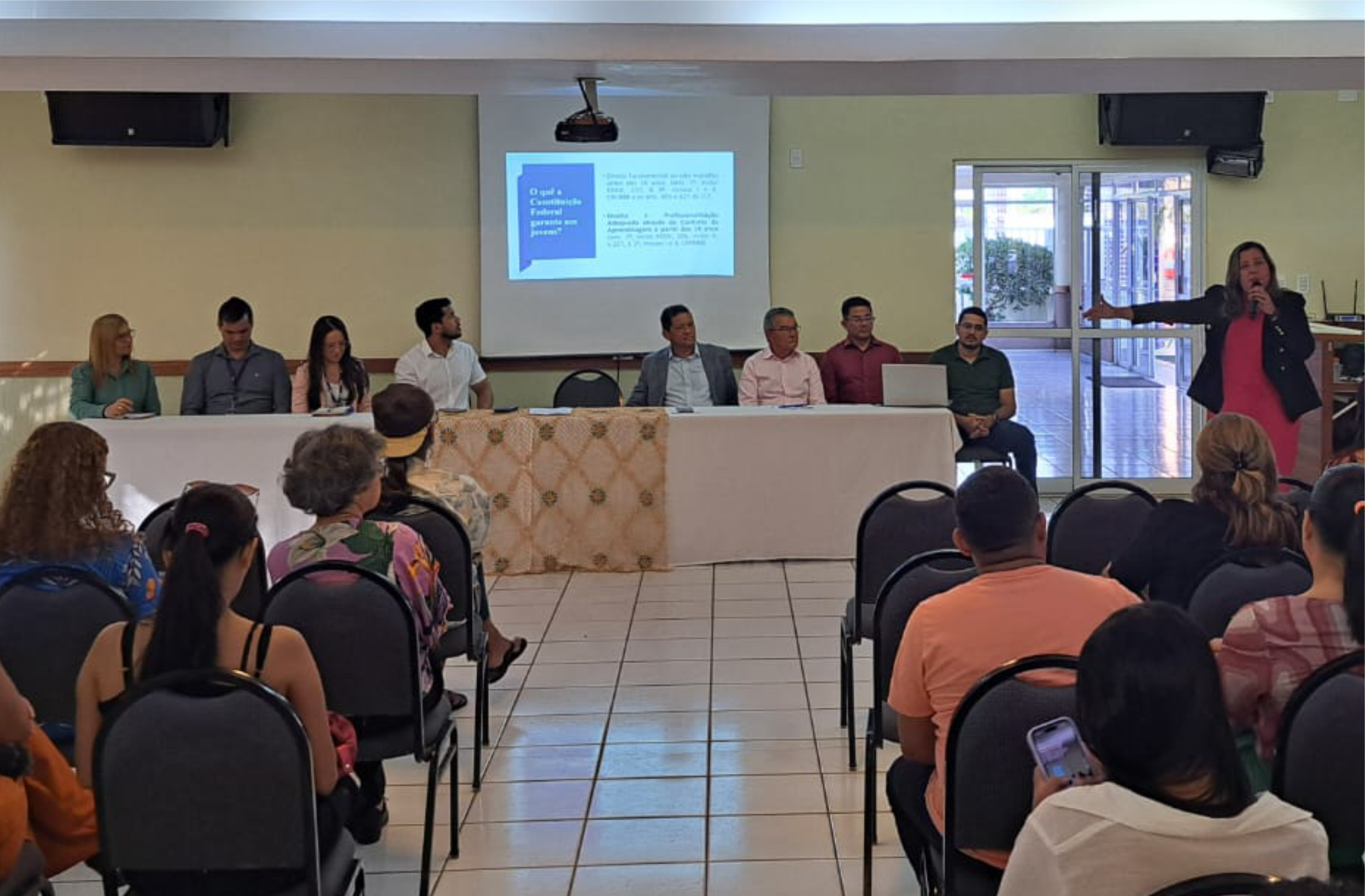 Foto de oito pessoas sentadas a uma mesa de frente para uma plateia. à direita, uma mulher de pé fala ao microfone, de frente para a plateia e apontando, com a mão direita, para tela de projeção, onde é exibida uma apresentação de slides. Plateia repleta de pessoas sentadas em cadeiras pretas.