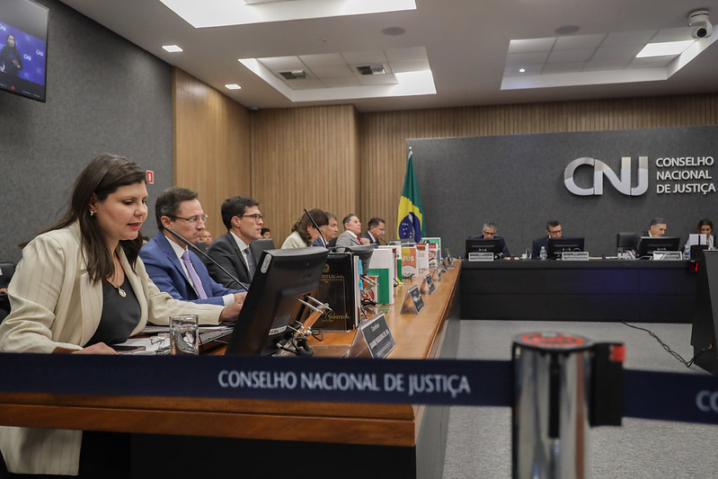 Foto de pessoas sentadas a uma mesa em forma de L, tendo à sua frente computadores e microfones. Ao fundo paredes amadeiradas e cinza escuro, onde aparece a logomarca do CNJ em metal prateado.