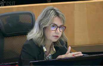 Imagem de uma mulher branca, sentada na cadeira de um plenário. Ela veste blusa estampada e um blazer  na cor preta. 