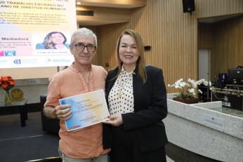 Foto mostra um homem com cabelos grisalhos e uma mulher branca no centro da imagem. Os dois seguram um certificado. Ao fundo um painel.