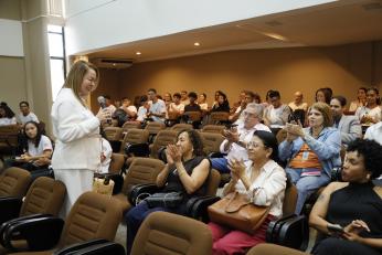 Pessoas sentadas em um auditório. Uma mulher branca se apresenta diante de todos.
