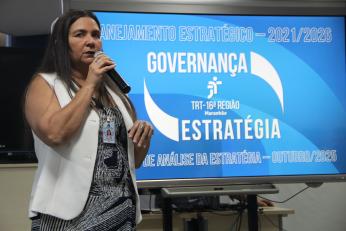 Foto de uma mulher falando ao microfone. Ao fundo um painel azul com letras brancas: Planejamento Estratégico 2021/2026. Governança Estratégica
