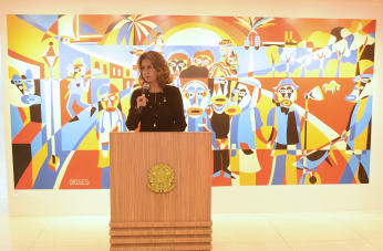 Foto mostra uma mulher em pé, atrás de um púlpito de madeira. Ao fundo uma pintura colorida retrata figuras que representam trabalhadores