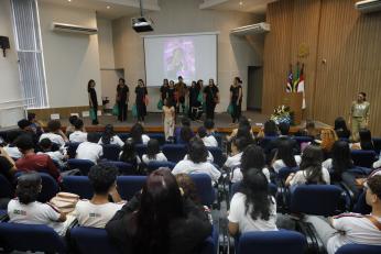 Foto mostra pessoas em apresentação no palco de um auditório. Outras pessoas estão sentadas nas cadeiras do auditório