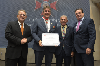 Foto mostra 4 homens em pé, um ao lado do outro. Todos usam termos escursos. Um deles segura nas duas mãos um certificado e usa um colar com uma medalha.