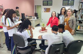 Foto mostra pessoas reunidas em volta de uma mesa. Ao fundo prateleiras com livros.
