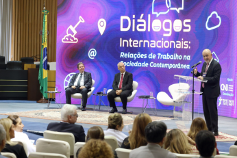 Foto mostra três homens usando terno escuro. Dois deles estão sentados e um em pé, falando ao microfone em um púlpito transparente. Atrás deles, telão de led exibe a identidade visual do evento. À frente deles, pessoas na plateia, sentadas em cadeiras brancas num auditório.