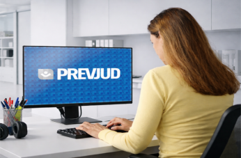 Foto de uma mulher clara de costas, usando blusa de mangas compridas amarela e cabelos longos. Ela está sentada a uma mesa, em frente a um computados, onde se lê na tela PREVJUD.