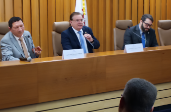 Foto de três homens claros, usando ternos e sentados em cadeiras marrons atrás de uma mesa de madeira. O do meio fala a um microfone, que segura com a mão esquerda.