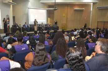 Foto mostra várioas pessoas sentadas em um auditório. Ao fundo, outras seis pessoas estão posicionadas no dispositivo de honra.