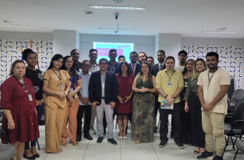 Foto mostra um grupo de pessoas em pé, são homens e mulheres. Todos estão em um auditório