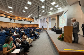 Foto mostra um homem em pé, atrás de um pulpito diante de uma platéia dentro de um auditório