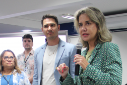 Foto de duas mulheres e dois homens. Em primeiro plano, à direita, mulher clara, de cabelos claros, usando blazer verde, fala ao microfone, que segura com a mão esquerda, e gesticula com a mão direita, olhando para a frente. Ao lado dela, as demais pessoas a observam falar.