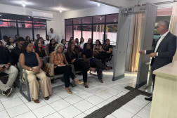 A foto mostra uma sala. Na parte direita, um homem em pé usa um terno escuro e camisa branca - sem gravata. Na esqueda vários homens e mulheres estão sentados. 