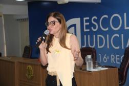 Imagem de uma mulher branca, usando uma camisa amarela. Ao fundo, uma parede azul com o nome na cor branca Escola Judicial. 