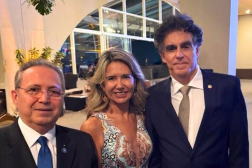 Foto mostra uma mulher posicionada ao lado de dois homens. Ambos estão com traje social.