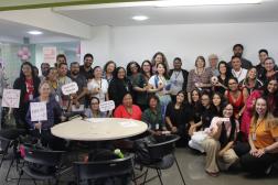 Foto mostra um grupo de pessoas, entre homens e mulheres reunidos em pé, e algumas pessoas agachadas. Alguns seguram nas mãos placas educativas sobre os cuidados contra o câncer de mama