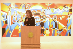 Foto mostra uma mulher em pé, atrás de um púlpito de madeira. Ao fundo uma pintura colorida retrata figuras que representam trabalhadores