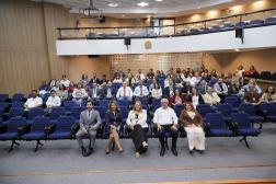 Foto mostra pessoas sentadas em cadeiras dentro de um auditório. Ao fundo, é possivel avistar um mesanino.