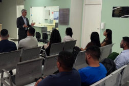 Foto mostra um homem, em pé usando terno escuro falando para pessoas que estão sentadas