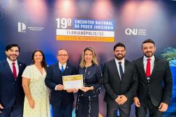 Foto mostra um grupo formado por seis pessoas. Todos estão em pé diante de um painel. Ao fundo a informação: 19º Encontro Nacional do Poder Judiciário