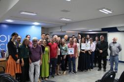 Foto mostra um grupo de pessoas. Todos estão em pé dentro de um auditório.