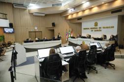 Foto mostra um plano geral de um plenário. Dez pessoas estão sentadas. Ao fundo as badeiras do Brasil, Maranhão e da Justiça do Trabalho.