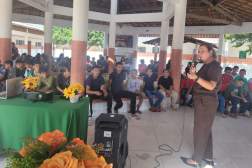Foto mostra um mulher em pé, realizando uma palestra para jovens estudantes. 