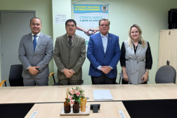 Foto mostra três homens e uma muher. Todos em pé, posicionados atrás de uma mesa de audiência