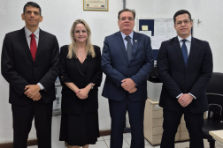 Foto mostra três homens e uma mulher. Todos estão em pé, um ao lado do outro em uma sala. Os homens usam terno escuro e a mulher um vestido longo e escuro.