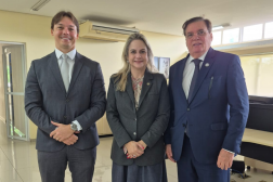 Foto mostra três pessoas, em pé. Sendo dois homens usando terno e uma mulher usando roupa social.