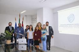 Foto mostra algumas pessoas em pé, posicionados em um auditório. Na frente delas uma mesa, com um jarro de planta e um notebook.