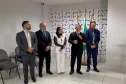 Foto mostra cinco pessoas em pé; Ao fundo uma parede branca com azulejos