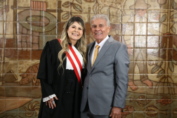 Foto mostra um homem e uma mulher. Ambos estão em pé. Ao fundo uma parede com azulejos marrons.
