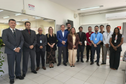 Foto mostra um grupo formado por homens e mulheres em pé. Um ao lado do outro. Ambos estão em uma sala.
