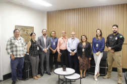 Foto mostra um grupo de pessoas em pé, dentro de uma sala. O grupo é formado por homens e mulheres.
