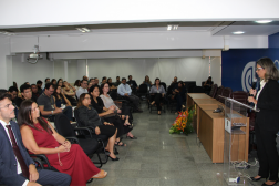 Foto mostra uma mulher em pé falando para uma platéia dentro de um auditório.
